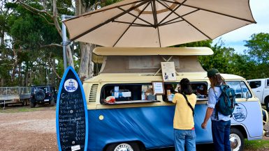 A blue VW Camper van with a cream roof sits in the middle of the frame. It is a coffee bar and a surfboard shaped notice board advertises the menu to the left. A large beige parasol sits above the van. A lady in yellow t-shirt and jeans, and a man in denim with long hair and a green baseball hat are ordering at the van window. Trees behind Margaret River shops