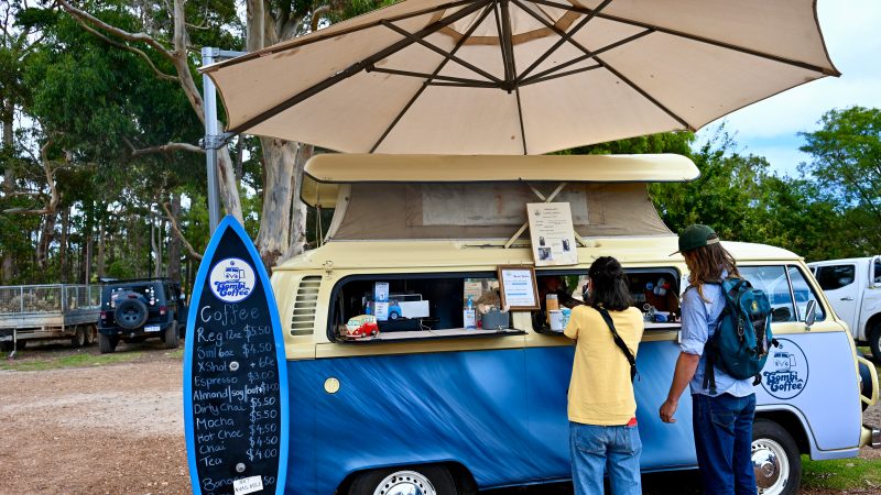 A blue VW Camper van with a cream roof sits in the middle of the frame. It is a coffee bar and a surfboard shaped notice board advertises the menu to the left. A large beige parasol sits above the van. A lady in yellow t-shirt and jeans, and a man in denim with long hair and a green baseball hat are ordering at the van window. Trees behind Margaret River shops