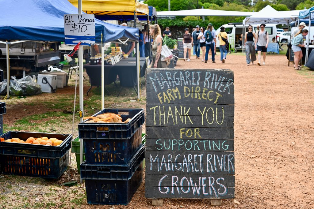 A black signpost sits on the floor in the centre of the frame advertising Margaret River Growers - thank you for supporting them. There are crates of produce to the left and market stalls behind. Gravel path to the right and people walking towards the camera