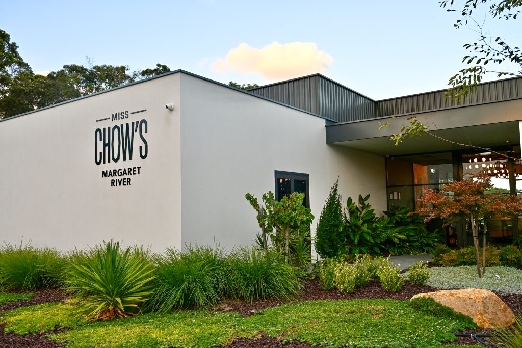 A white new build, single-storey building sits to the left of the frame with green lawn and plants in front. It says 'Miss Chow's Margaret River' on the side on black writing. Trees and blue sky above. Glass doorway to the right