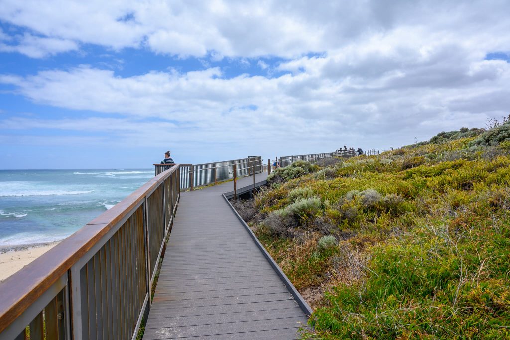 View along a boardwalk leading to the middle of the horizon. Grass land to the right. Turquoise ocean to the left. Cloudy sky above