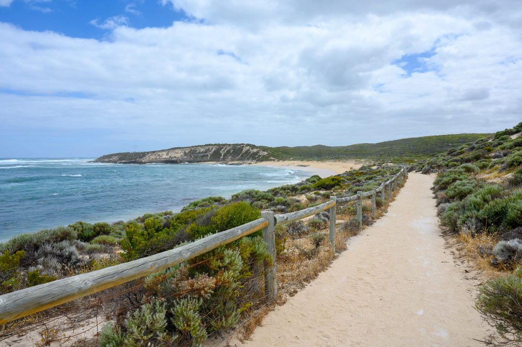 View along a path to the right hand side, wooden barrier to the left and shrubland beyond. Turquoise sea to the left. Cloudy blue sky above
