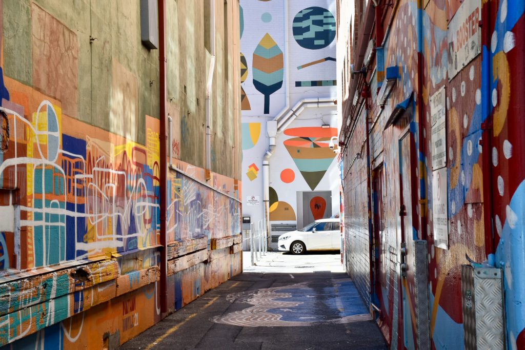 Alleyway looking down to a street at the end. The walls are painted with murals with geometric shapes, as is the wall of the building at the end. There is a white car parked at the bottom of the alley- illusion of being in a painted box