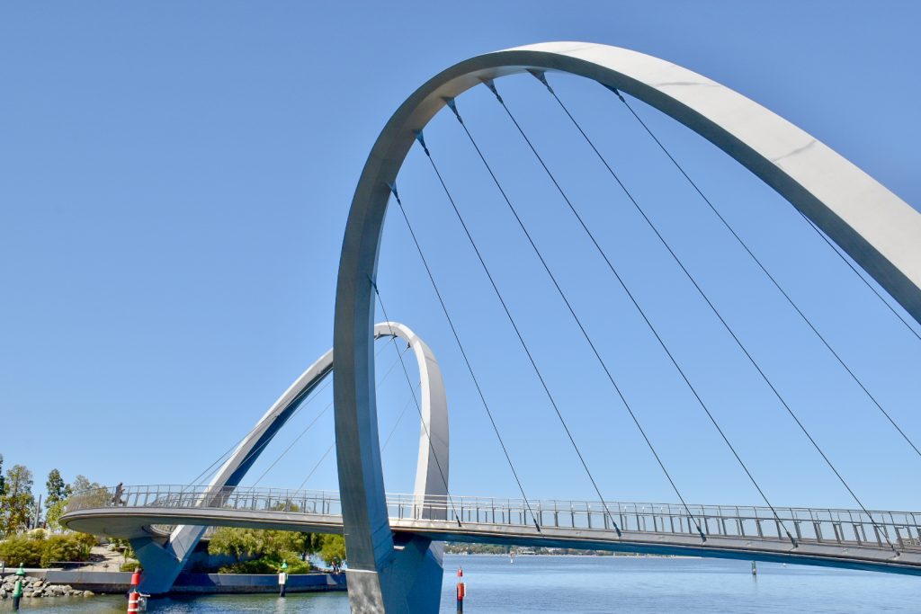 Pale blue sky with a metal bridge reaching from the bottom right of photo across to bottom left. The bridge as two arches, one of which leans to the middle of the photo