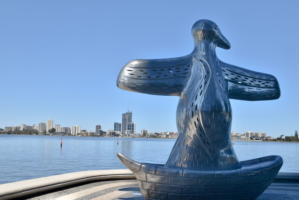 Statue of a bird like metal structure with wings open, sitting on waterside with buildings on far side of the river