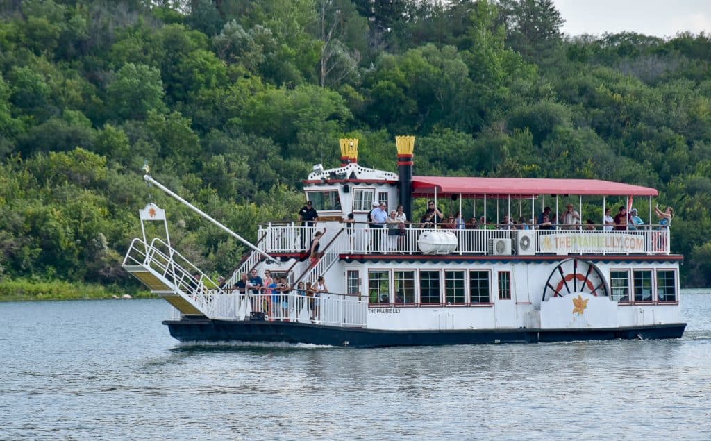 Flat grey waters of river in bottom half of photo. Thick body of green trees in background. On the water is a white paddle boat style boat. It has a red canopy offering shade on the top deck and a bottom deck with windows, a wheel on side of boat and at the front a raised gangplank. There are a number of passengers on the boat admiring the views