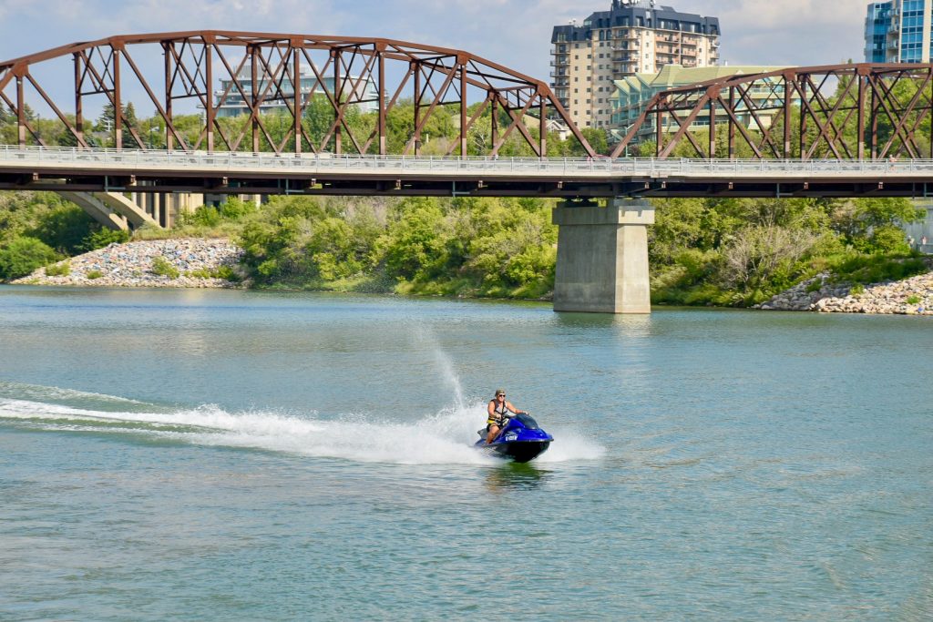Photo of a grey-blue river with a metal bridge spanning the river. Green shrubs and building on the far river bank. In the centre of the river and frame is a man riding a blue jet ski which is shooting a jet of water behind
