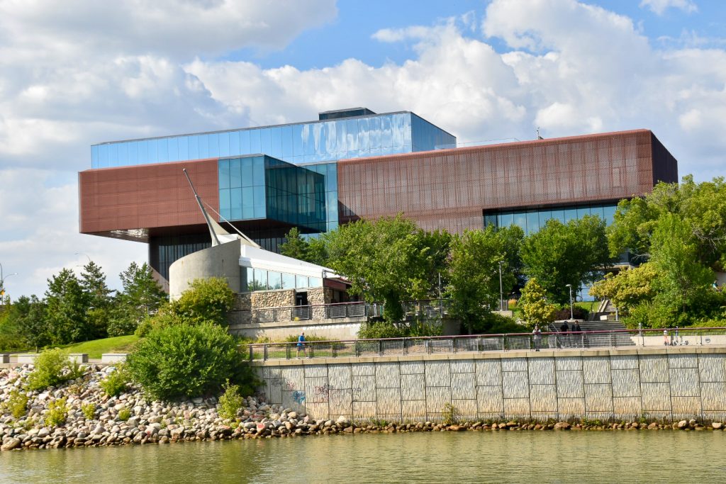 Flat green river water at bottom of photo. Image taken from the river looking at a very modern building on the bank - the building appears like a number of blocks piled on top of each other. Cloudy blue sky in background