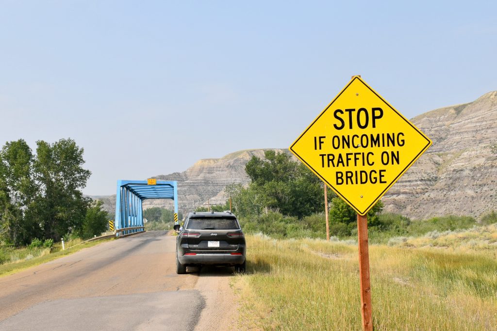 Photo of a yellow road sign to the right telling cars to 'STOP if oncoming traffic on bridge', black car sitting in the road with a blue bridge behind