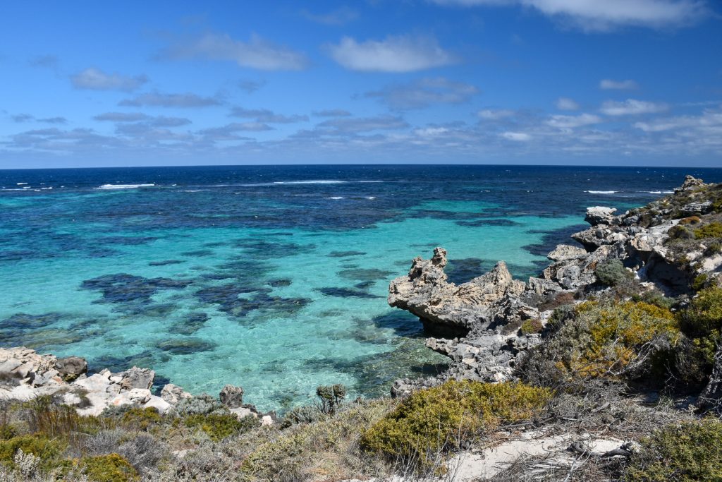 Blue sky with some white cloud in top third. Foreground has grey rocky shoreline with some shrubs. Beyond the rocky shoreline is a clear turquoise sea with patches of darker blue
