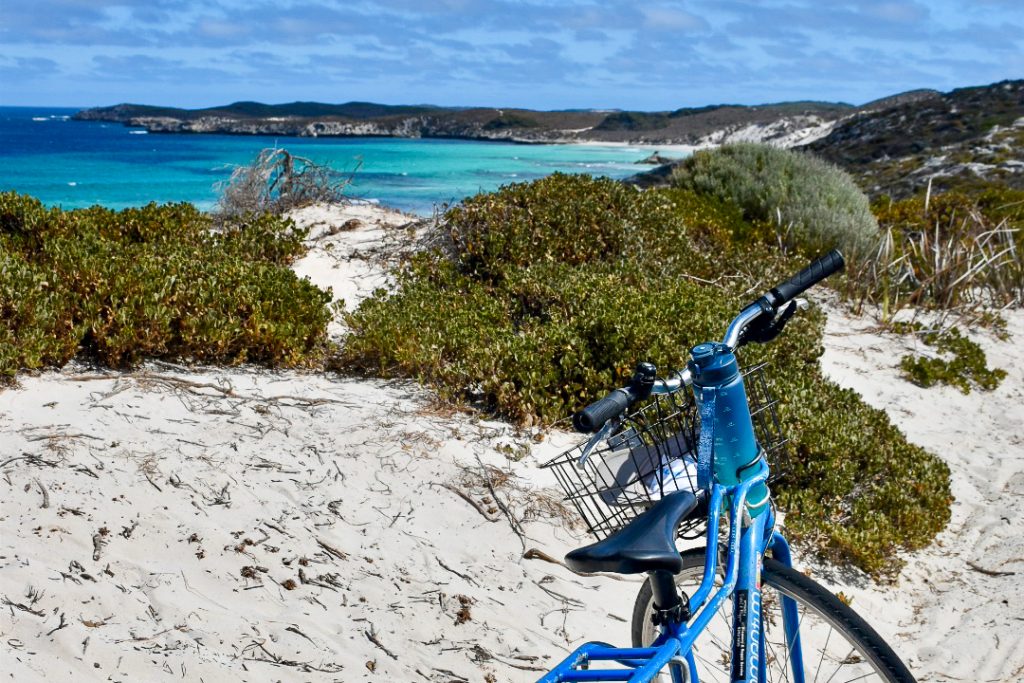 White sand in foreground, green shrubs on the cliff overlooking pretty turquoise water in a curved bay with green headland and blue kay beyond. A blue bicycle with a wire basket on the front is parked in the bottom right hand side of the frame