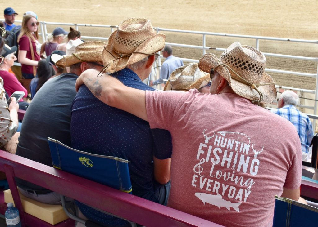 The backs of two men in cowboy hats, sitting in a grandstand overlooking a rodeo arena. The man on the right has worn a pink t-shirt which says Huntin', Fishing and Loving life. He has his arm around the man to his left