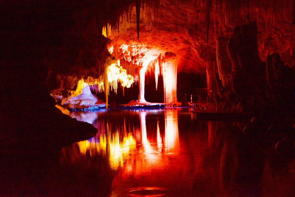 Dark scene where some rocks formations are illuminated in red and yellow, and reflected in a shiny surface below