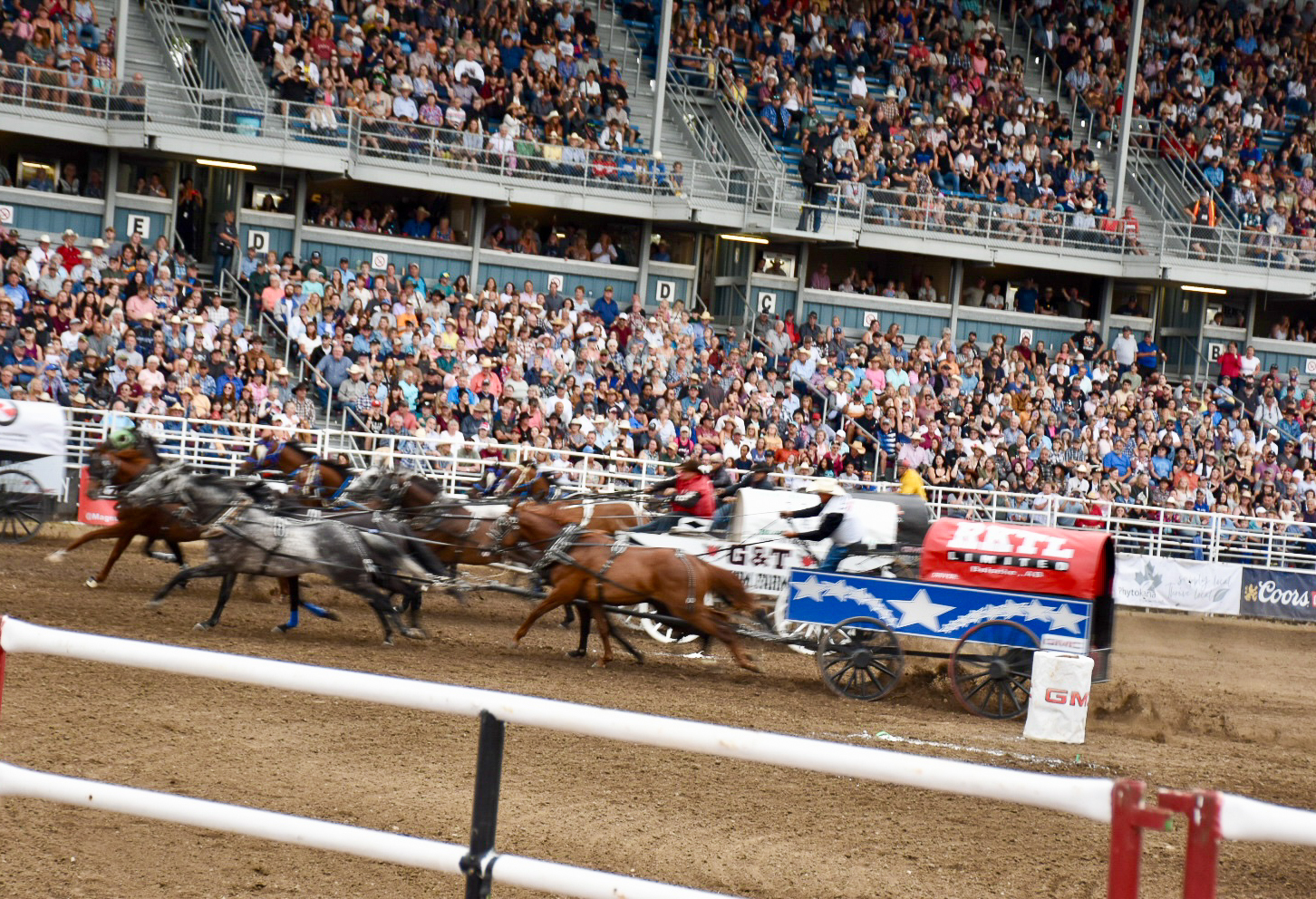 View over white railings into an arena where some colourful wagons are racing past.  Full standium of visitors in background