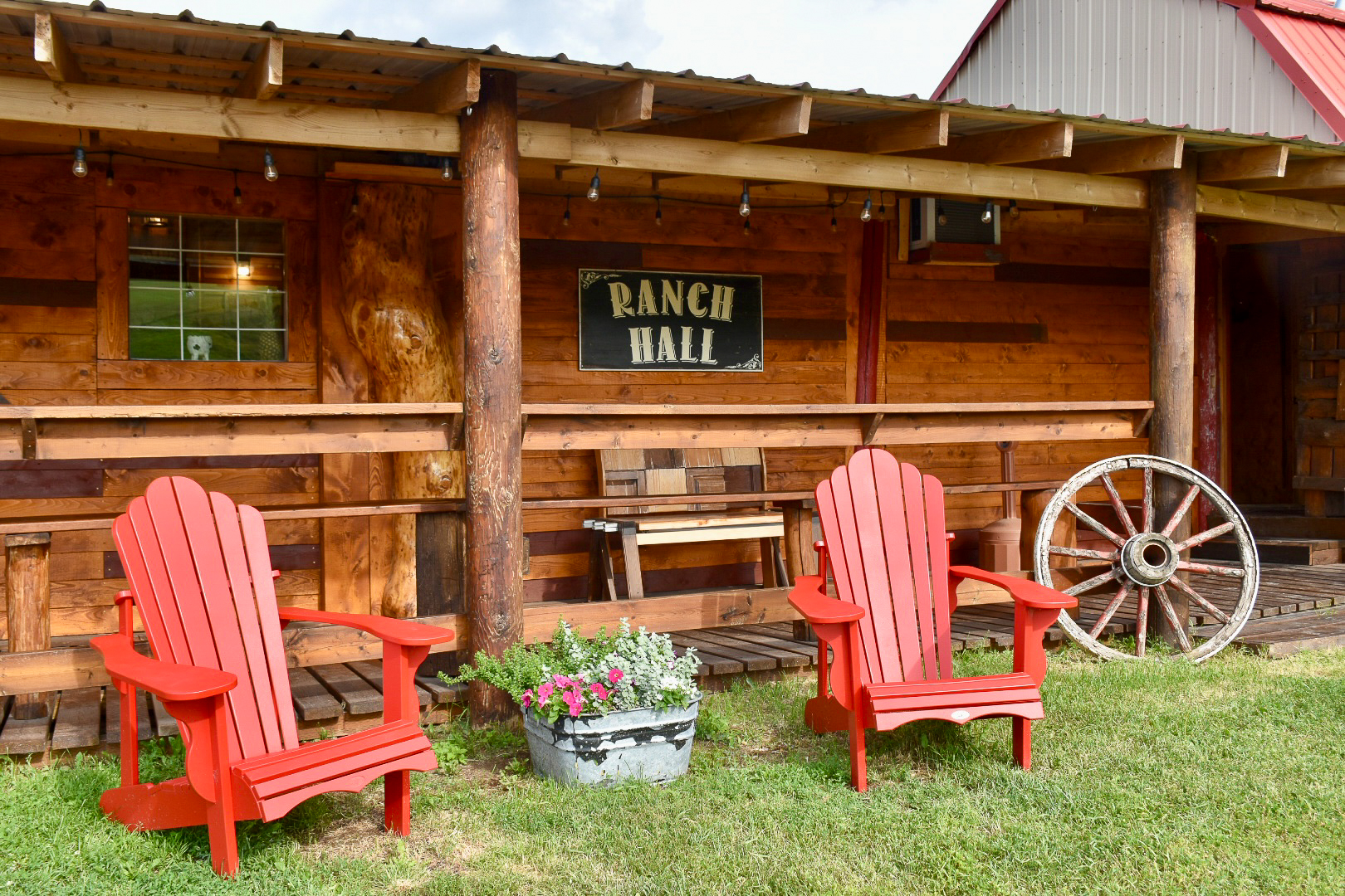 Two red aridonack chairs sitting outside a long brown wooden barn with the words Ranch Hall on the wall