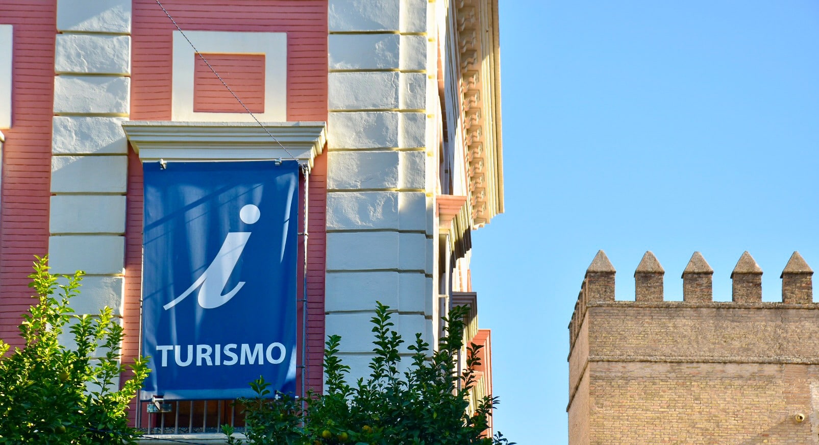 Left hand side of photo shows the side of a white brick building with pink panels. From the window hangs a blue banner which says 'Turismo' with a large 'I' on it for information. The right hand side looks down the front of the building to a pale blue sky and the turrets of a castle still building in bottom right of frame