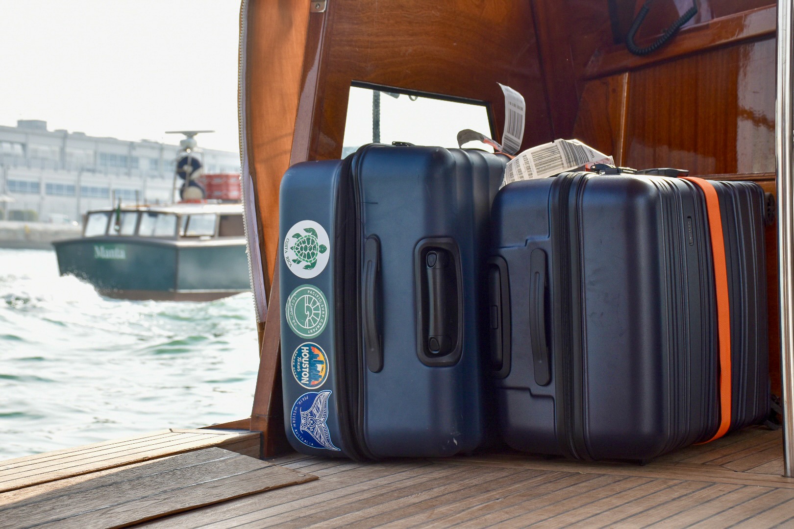 Two navy blue suitcases lie on their sides on the floor of a wooden boat. The left suitcase is slightly bigger and features 4 stickers (one says Houston on it). The right hand suitcase has an orange strap. Both cases have their white airline tags showing on the top. To the left hand side of the boat, you can see the water through the side and a boat sailing past