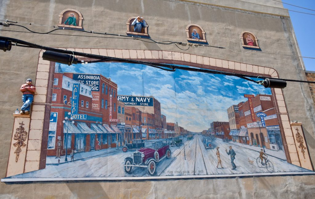 Mural of old Canadian street painted on side of building, around the mural are some 3D figures - one stands to the side and one is looking out of a window above