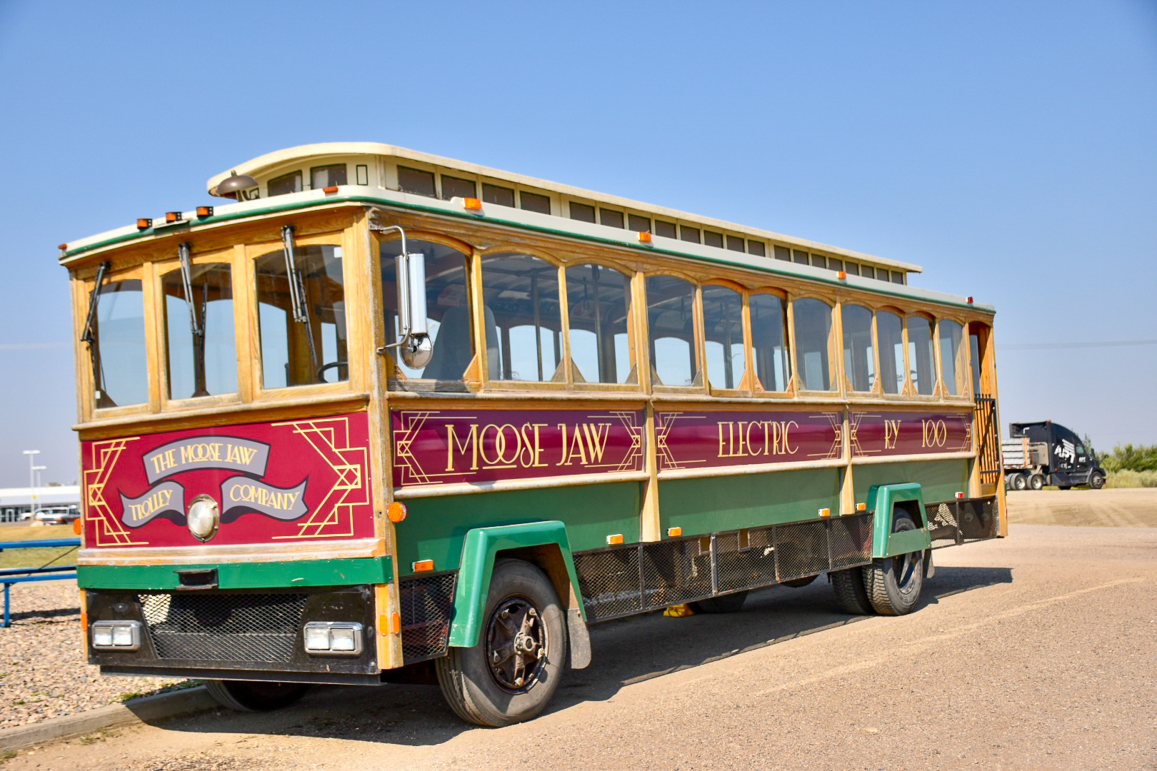 Old fashioned trolley bus parked on road against a pale blue sky.  The trolley bus is green at the bottom, with red stripe around middle and yellow top.  Moose Jaw Trolly painted on bus