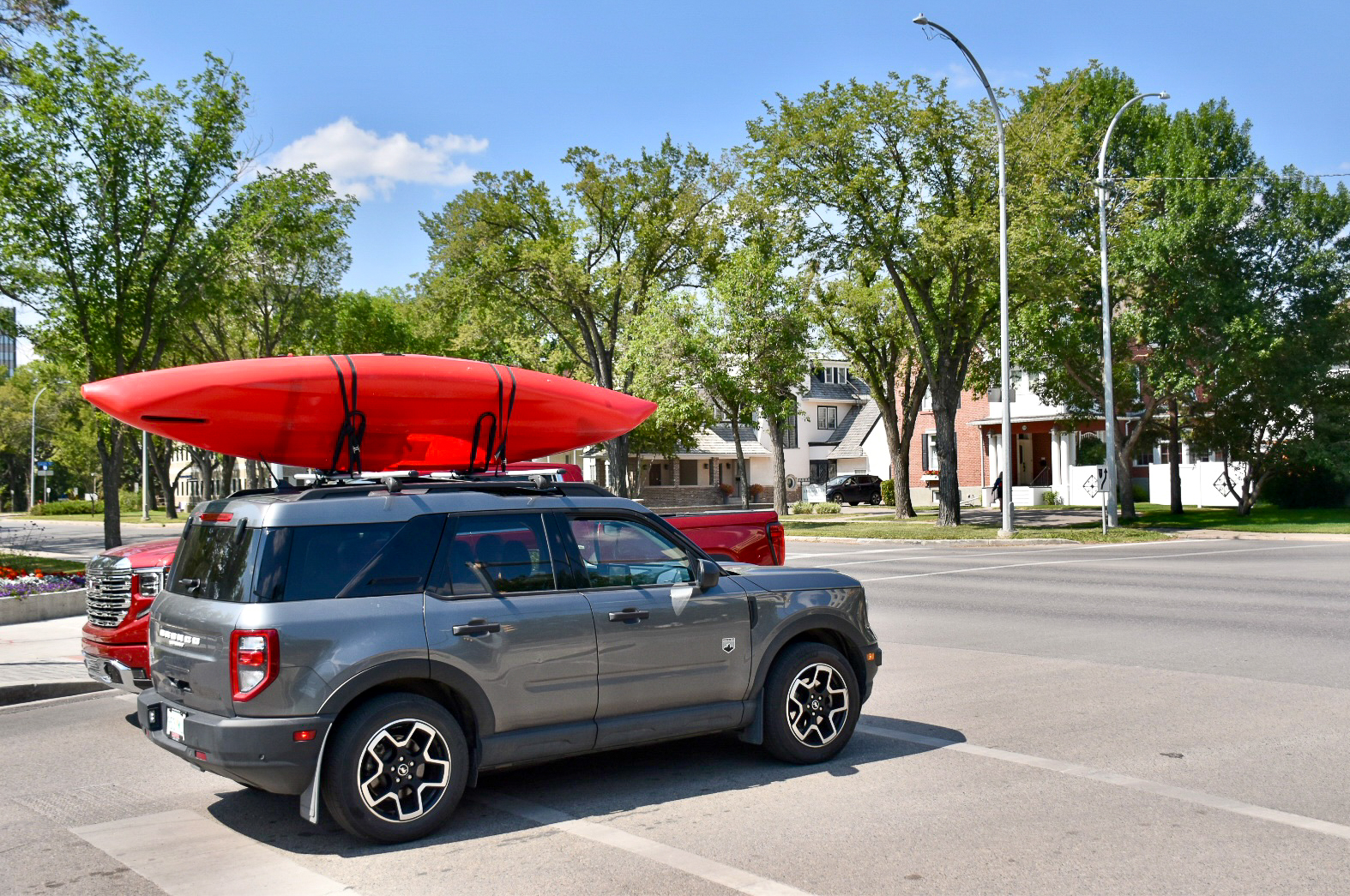 Silver SUV car with red kayak on its roof. Car is coming into photo from bottom left corner, waiting to move into the centre of road. Trees on the other side of road and hint of blue sky