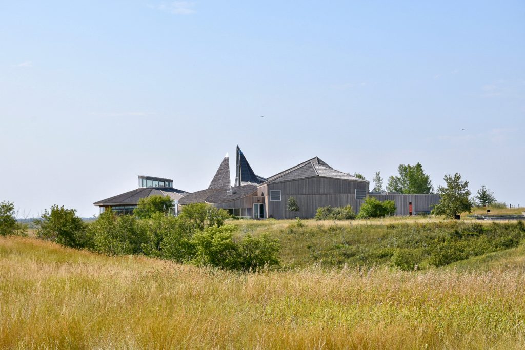 Pale blue sky at top of photo. Grasslands in bottom half of photo with hints at a valley just before the midpoint. On the horizon is a series of modern buildings with some pointed roof and glass features. Made from wood.