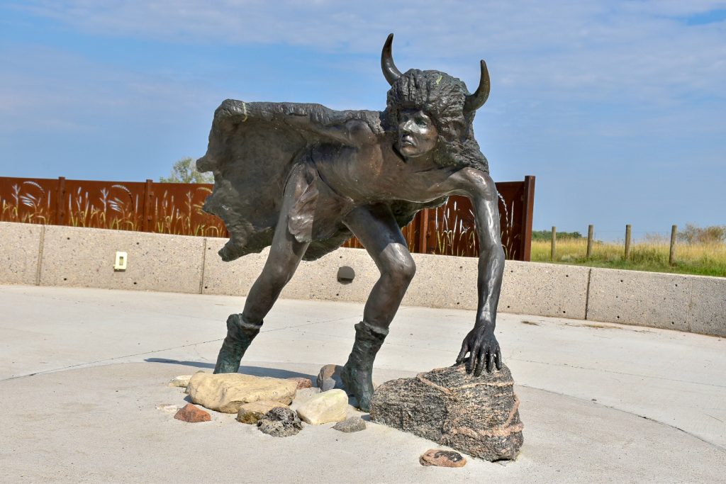 Statue of a Native American Calf Runner looking towards the camera with a plain concrete floor and building behind in distance