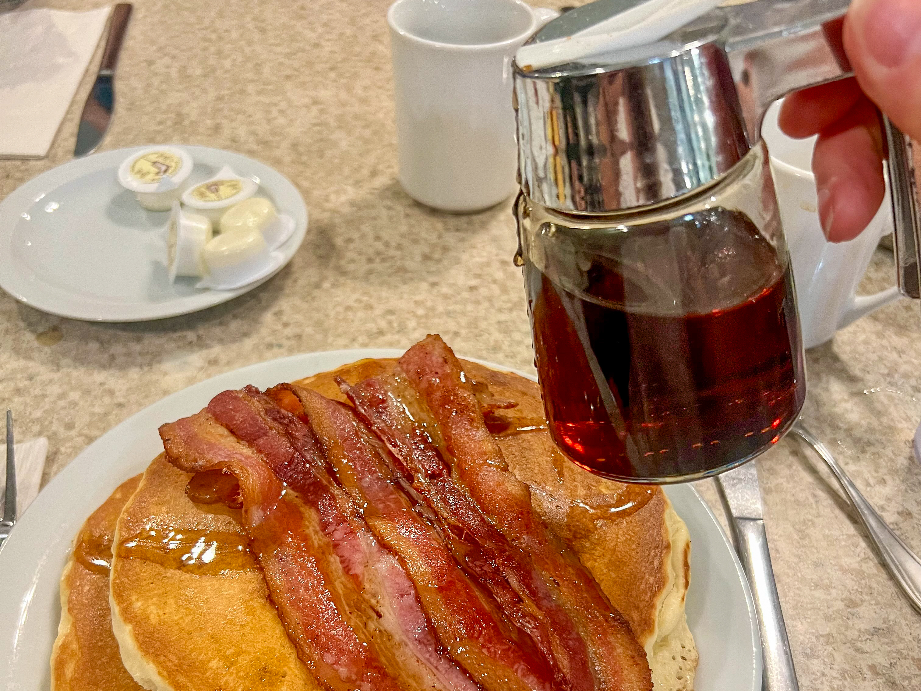 Cafe table shot of pancake stack with bacon and jar of maple syrup about to be poured.