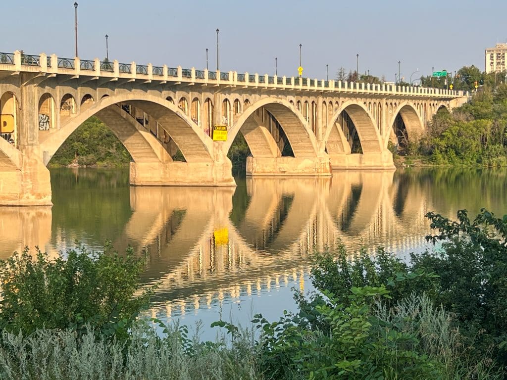 Stone bridge over the South Saskatchewan River in Saskatoon at sunset, glowing gold with its full reflection in the still water below.