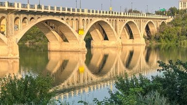 Stone bridge over the South Saskatchewan River in Saskatoon at sunset, glowing gold with its full reflection in the still water below.