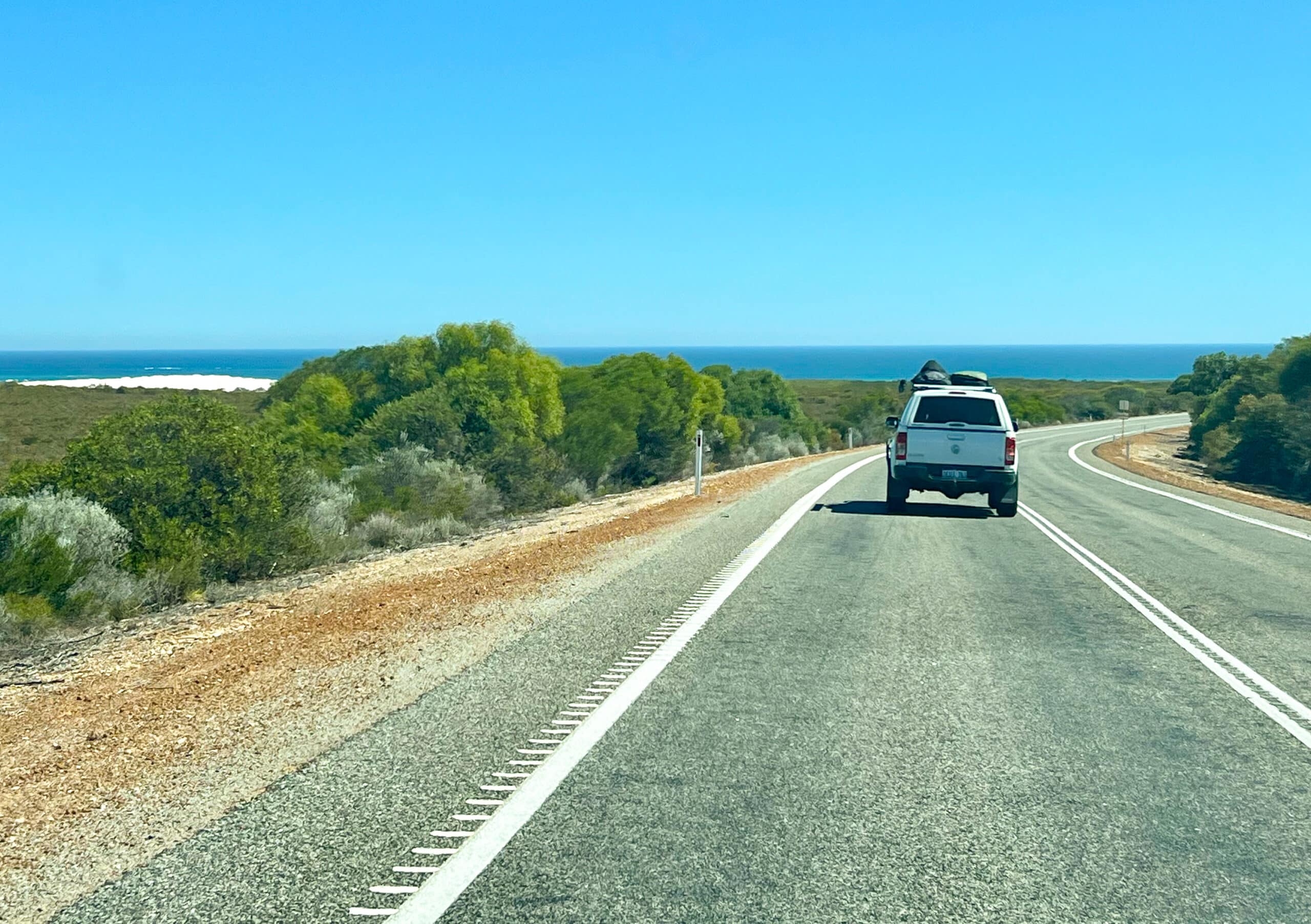 A grey tarmac road with white lines runs from the front of the frame and curves around to the right at the middle. Green shrubs on either side of the road, some white dunes on the horizon to the left beyond the shrubs, blue sea on horizon. Pale blue sky