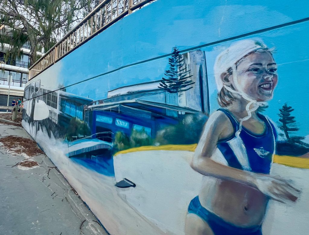 Image taken up an alleyway so the right hand wall fills the frame from left hand side diminishing to the middle left hand side. Wall is decorated by a mural of a young girl in blue swim wear and white swim cap running towards sea with white and yellow surf board. Second surfer in background and blue buildings in-between the two surfers