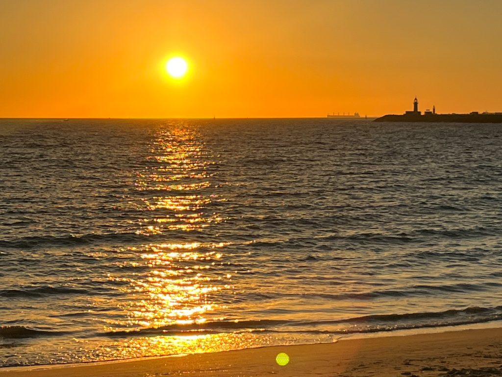 Golden sunset photo. Sun is still quite high on the horizon at top of the photo, casting its golden glow across the water below, down to the beach in the foreground. Silhouetted lighthouse on the horizon