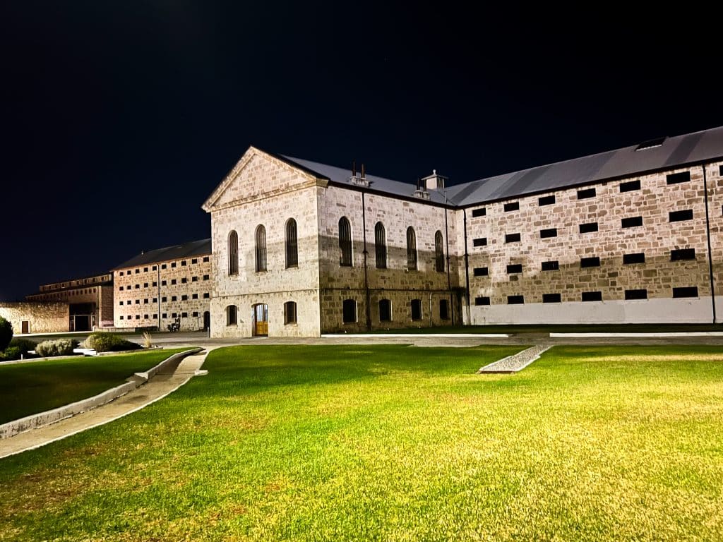 Night time photo - black sky. Brick prison building across the middle of the photo. Green grass illuminated by the spotlights in foreground