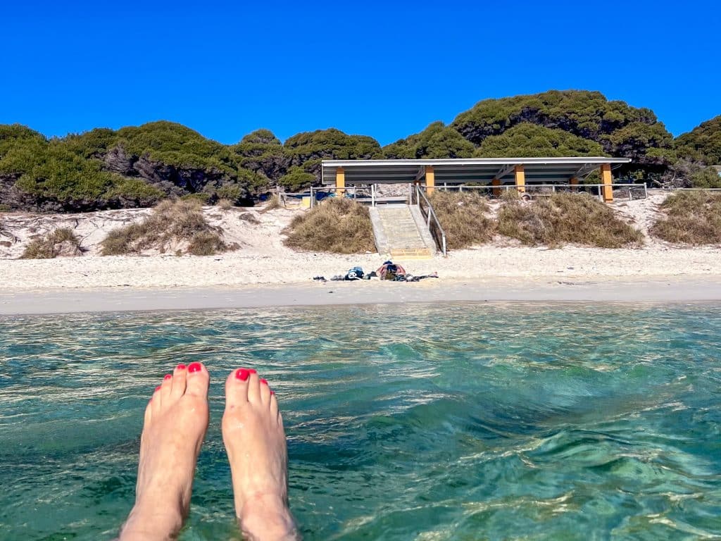 Pair of feet with red toe nails floating in clear turquoise water. View back to a white sandy beach and simple pavilion with trees in the background. Dark blue sky with no cloud