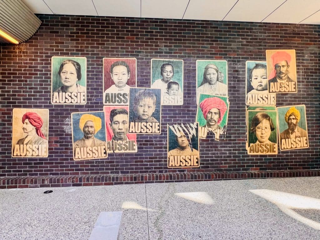 Posters on a brown brick wall with pavement in front. Each poster features a photograph of a person's face with enhanced colour. Each poster reads Aussie