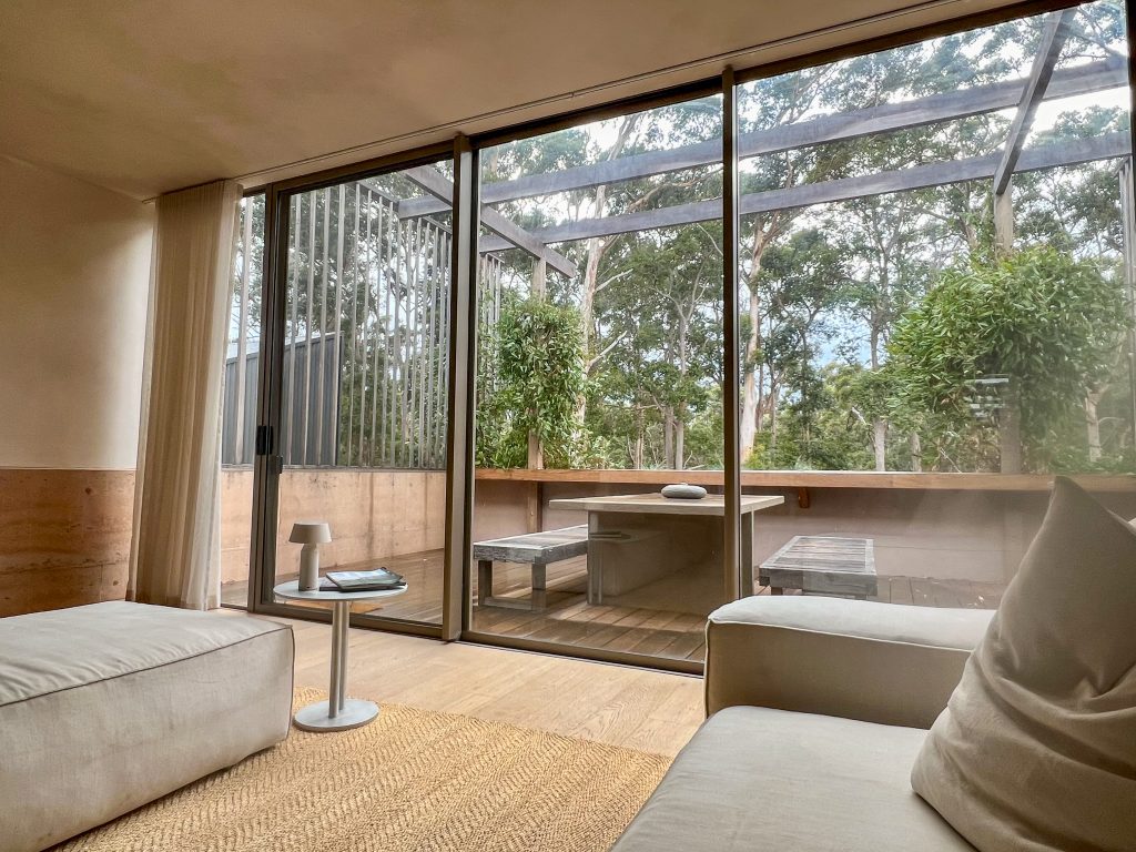 View across a lounge with beige sofas to floor to ceiling windows out to tall trees beyond