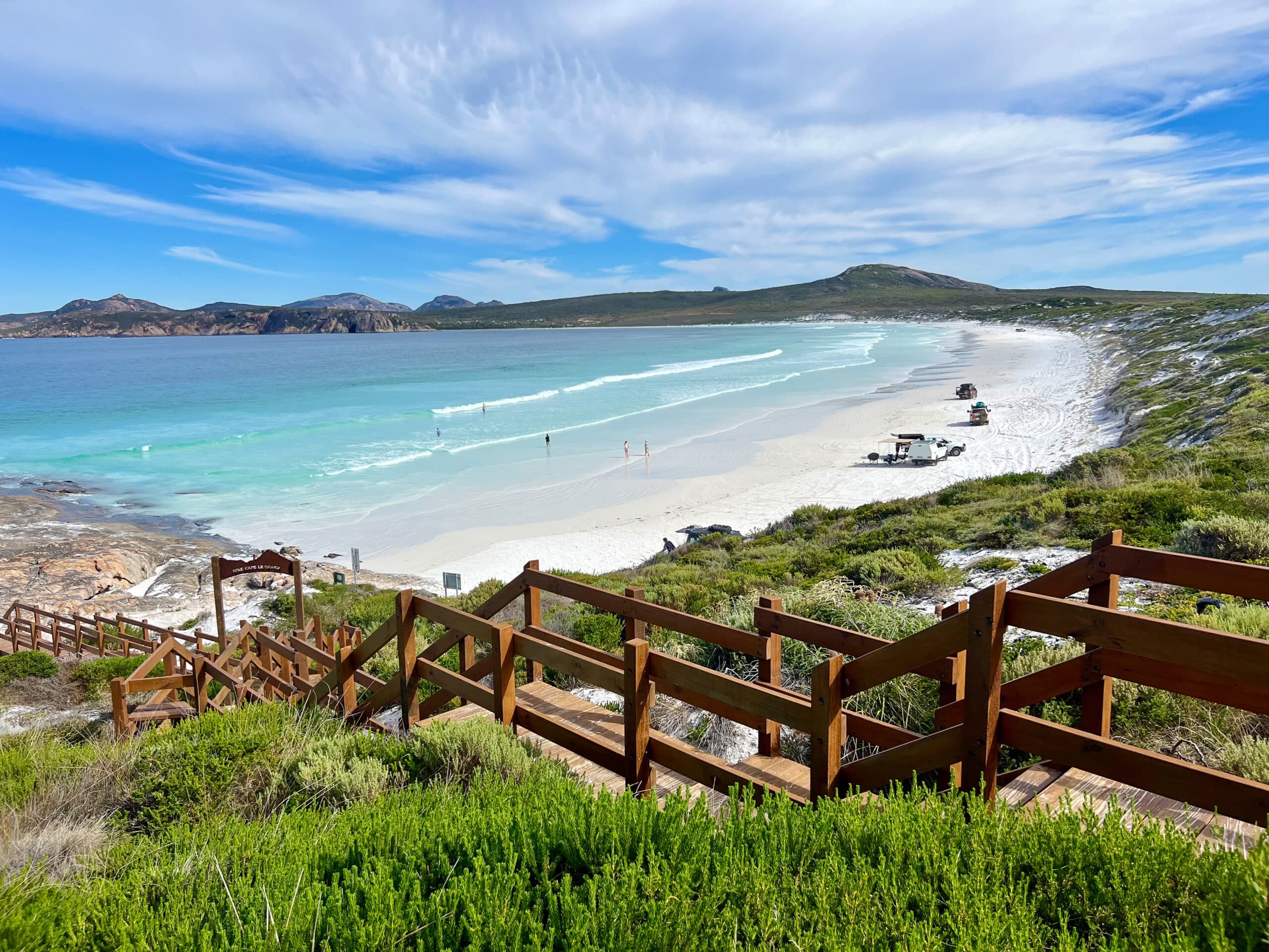 Beach landscape with green shrubs in the foreground and wooden staircase leading down to a white beach to the right of a pale turquoise ocean. You can see land in the distance creating a curved beach