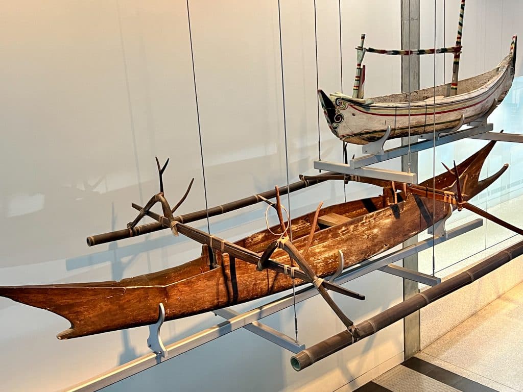 Two wooden boats exhibited in museum. The front boat is made of brown wood and is like an old fashioned canoe. The back boat is similar but smaller and white wood. They are indigenous boats