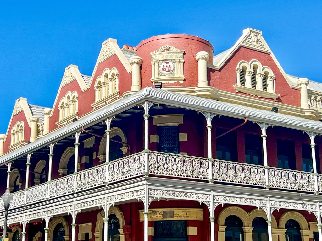 Detail on an old fashioned Gold Rush building with a dark pink roof with white plaster work, above white painted iron work balconies and verandas. Blue Sky