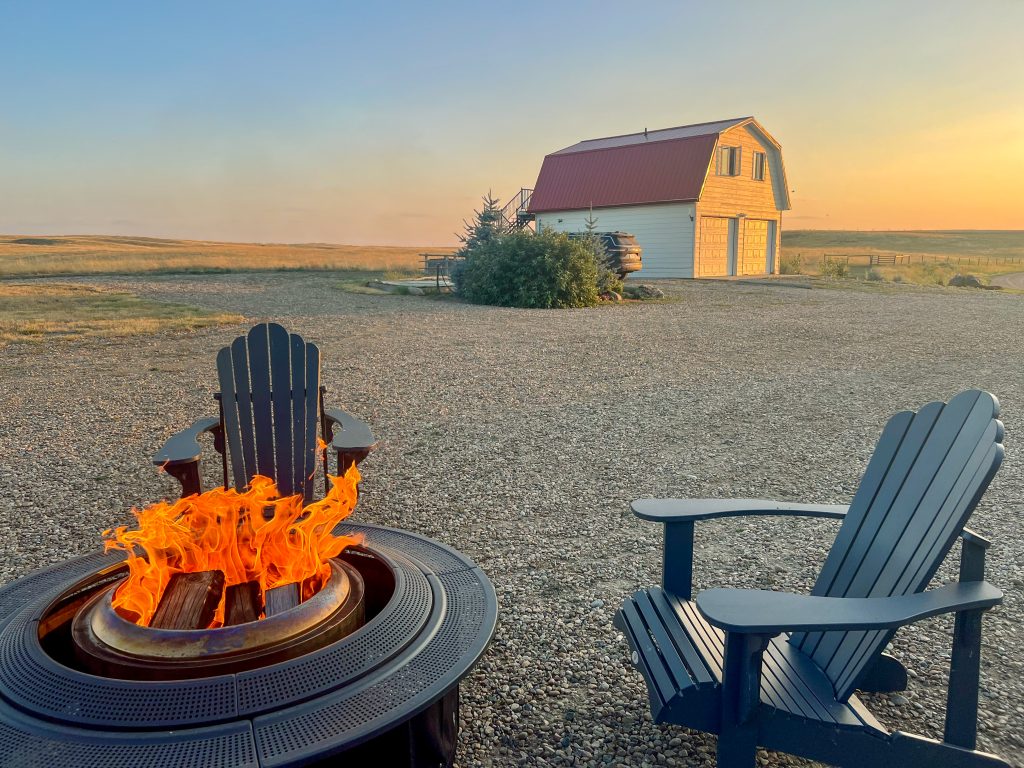 Fire pit and green chair in the foreground. White barn with red roof in background against a setting sun