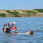 Person floating in a blue lake with small hills in the background. The person is reading a magazine whilst floating