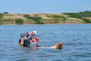 Person floating in a blue lake with small hills in the background. The person is reading a magazine whilst floating