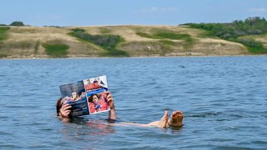 Person floating in a blue lake with small hills in the background. The person is reading a magazine whilst floating