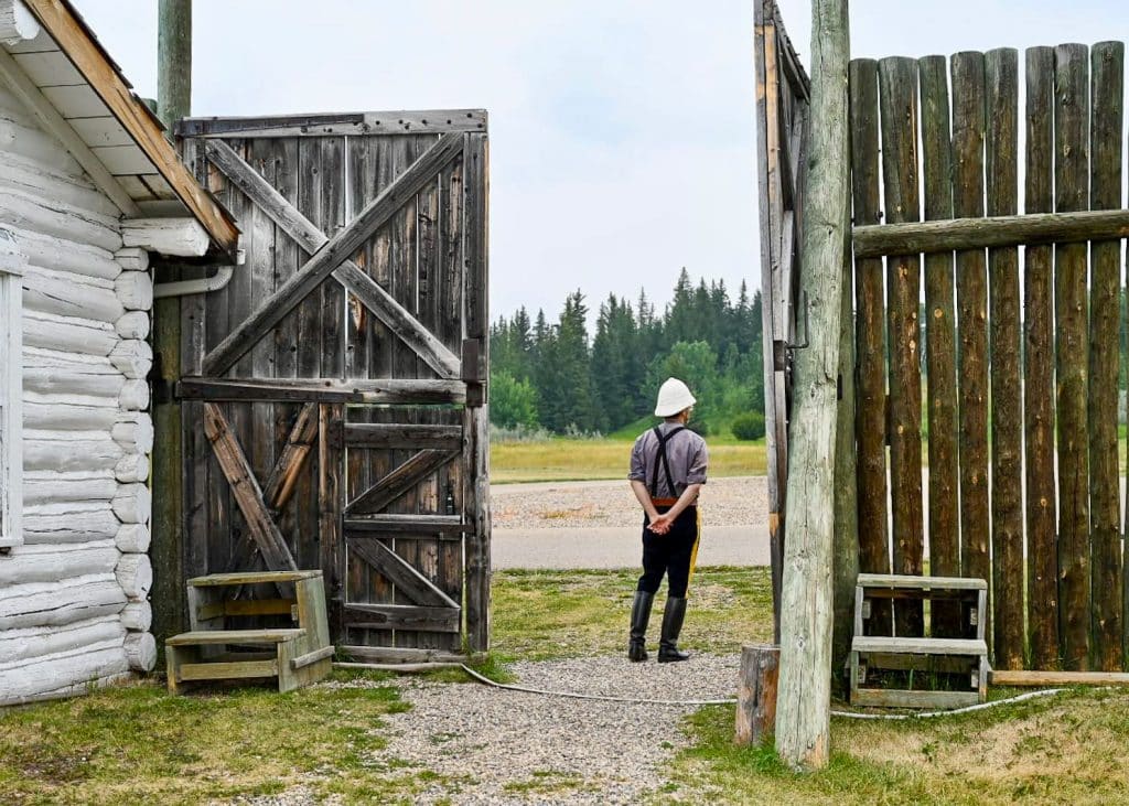 Tall wooden fence (twice as high as a 6 foot man) with open wooden door. White wooden casing to the left of the photo. A man in old fashioned Mountie uniform and white hard hard looks away from the camera towards some trees in the distance