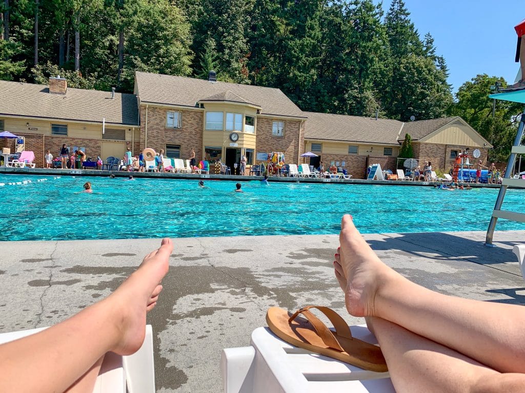 Outdoor swimming pool with row of brown buildings behind the pool and fir trees behind the buildings. Foreground shows two sets of feet belonging to people relaxing on sunbeams