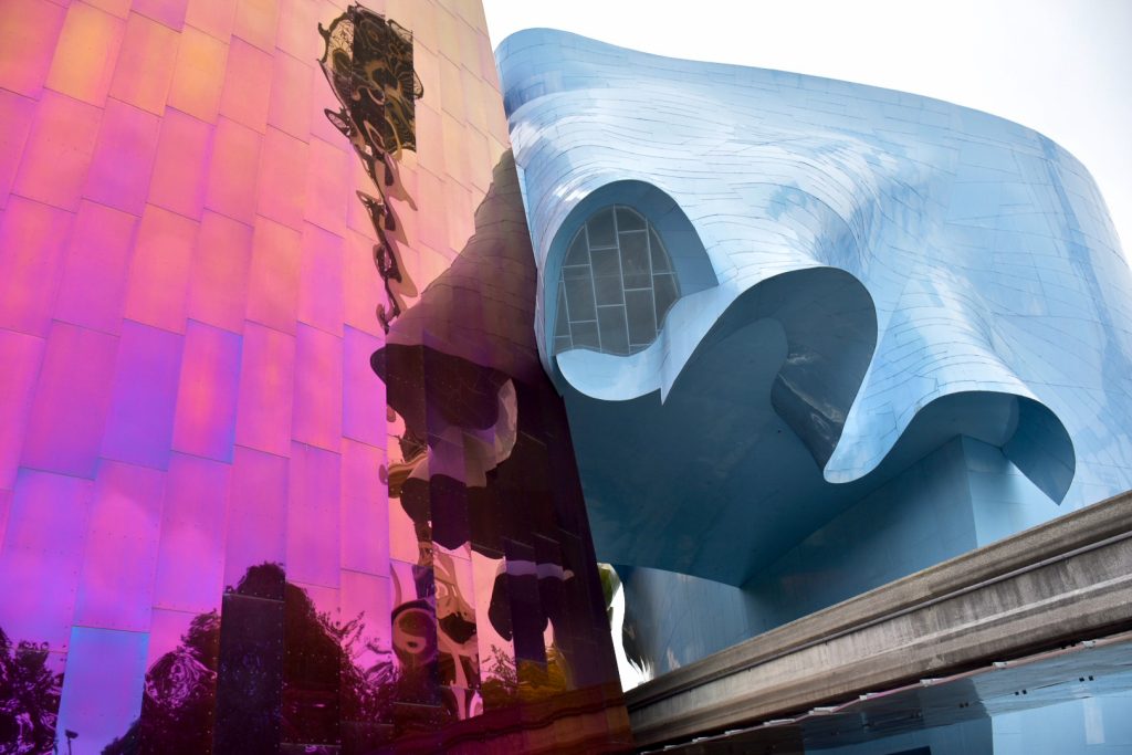 Unusual coloured and shaped modern building. Left half of building is covered in pink, purple, orange shiny metallic panels. The right hand side of the building has wavy edges across the middle like a wave