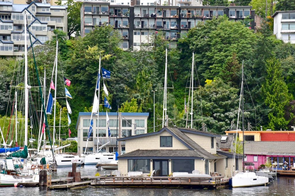 Photo of a large houseboat (beige and brown) on a platform on the water at the bottom of Frame surrounding by a number of sailing boats. Behind is a row of evergreen trees and behind that several blocks of flats