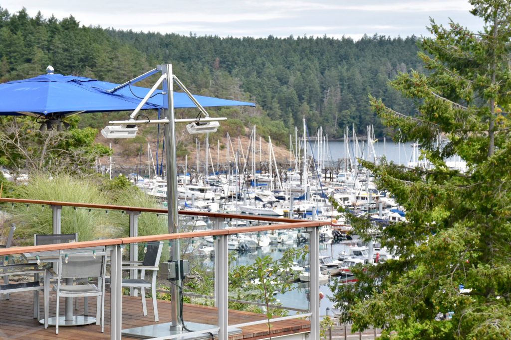 Foreground has the corner of a restaurant terrace with seats and blue awning overhead, sailing boats parked up in a marina behind with evergreen trees on horizon