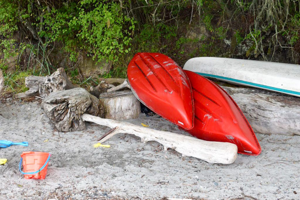 Upturned canoes - 2 red and 1 white - on sandy beach with driftwood and an orange bucket and spade. Green trees behind