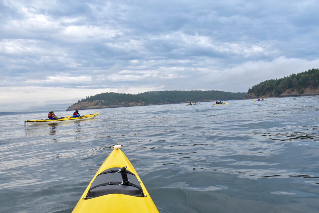 Flat grey waters and grey cloudy sky. Foreground shows the bow of a yellow kayak. There is a strip of tree covered land on the horizon and a second yellow kayak (double) ahead to the left on the water. In the distance are 3 other double kayaks